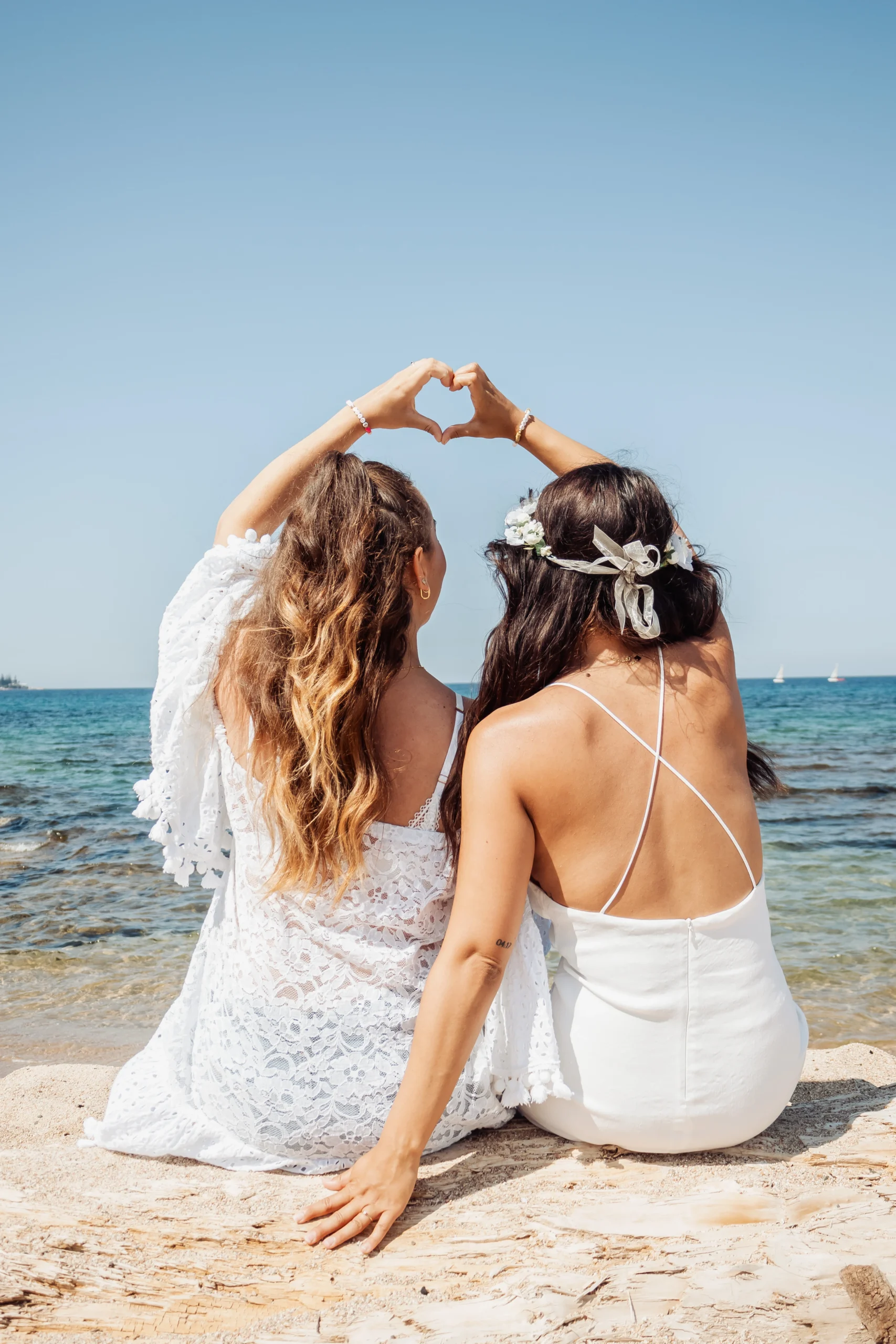 evjf-ajaccio-coeur-dos-bord-de-mer-plage Deux femmes assises sur la plage à Ajaccio lors d’un EVJF, de dos, formant un cœur avec leurs bras face à la mer, ambiance poétique et complice.