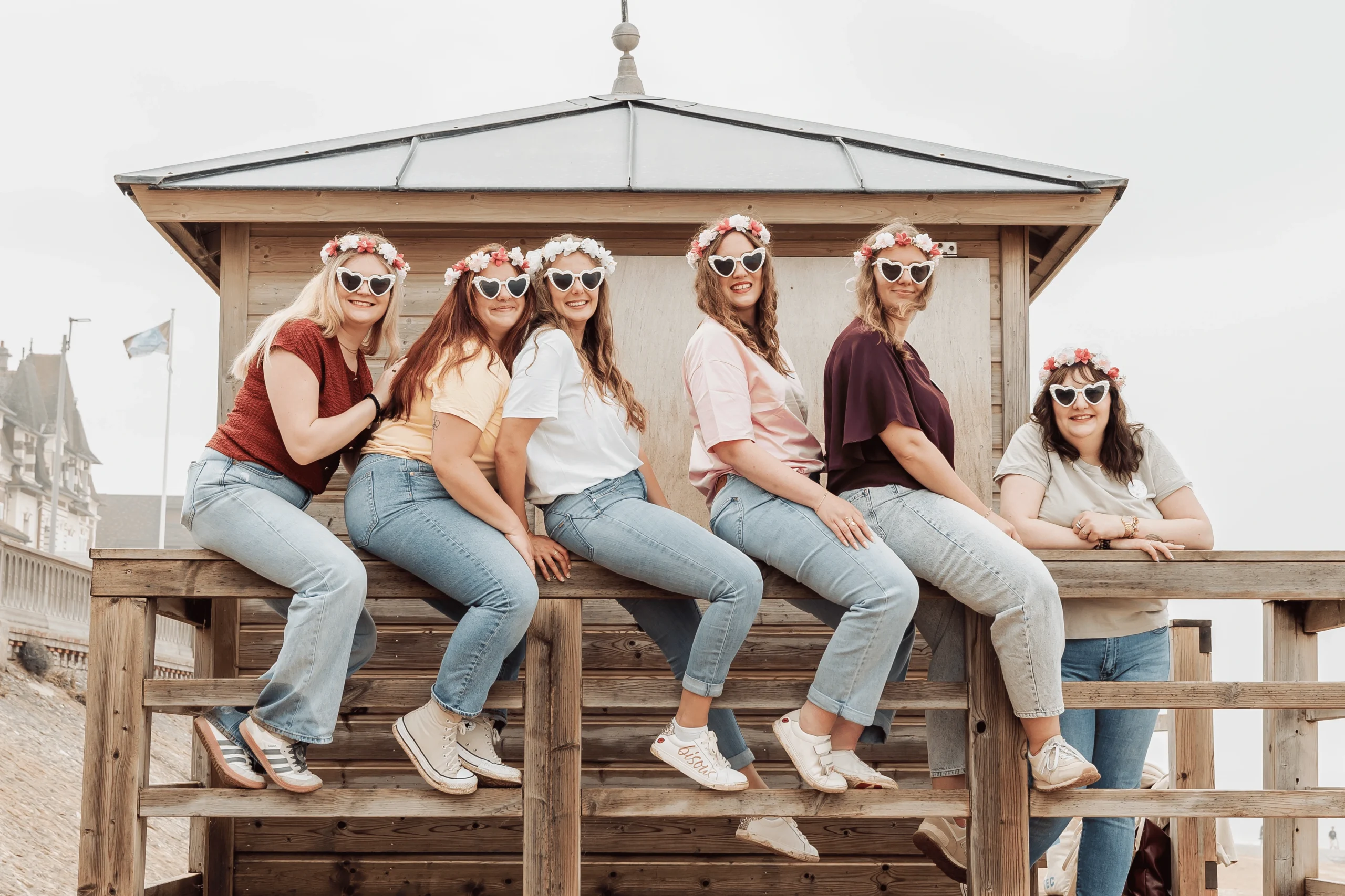 Photo EVJF à Cabourg avec le groupe assis sur une cabane en bois face à la plage, lunettes cœur et couronnes de fleurs. Ambiance bord de mer et fun.