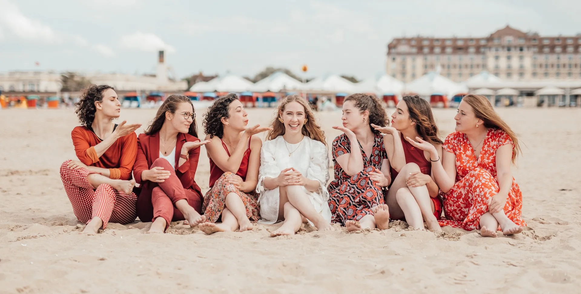 EVJF à Deauville – Moment tendre entre amies, bisous soufflés vers la mariée assise au centre, fond de plage