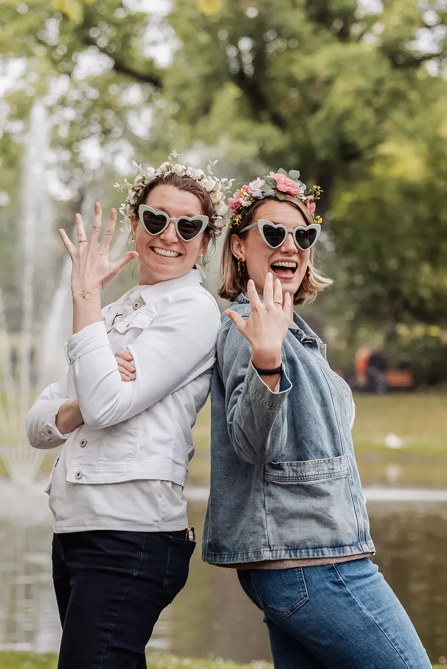 EVJF à Nantes – Deux amies complices posent avec lunettes cœur et couronnes de fleurs Deux femmes souriantes portant des lunettes en forme de cœur et des couronnes de fleurs posent dos à dos devant un étang dans un parc à Nantes.