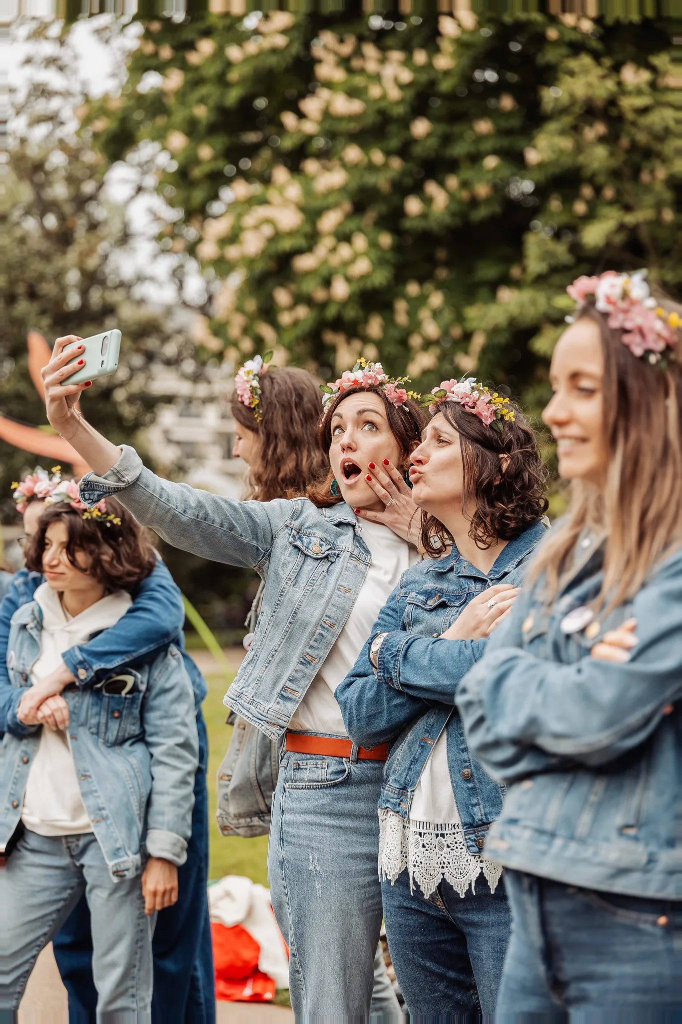 EVJF à Nantes – Moment fun entre amies avec selfies et couronnes fleuries Un groupe de femmes en veste en jean avec des couronnes fleuries, prenant un selfie drôle pendant un EVJF dans un parc de Nantes.