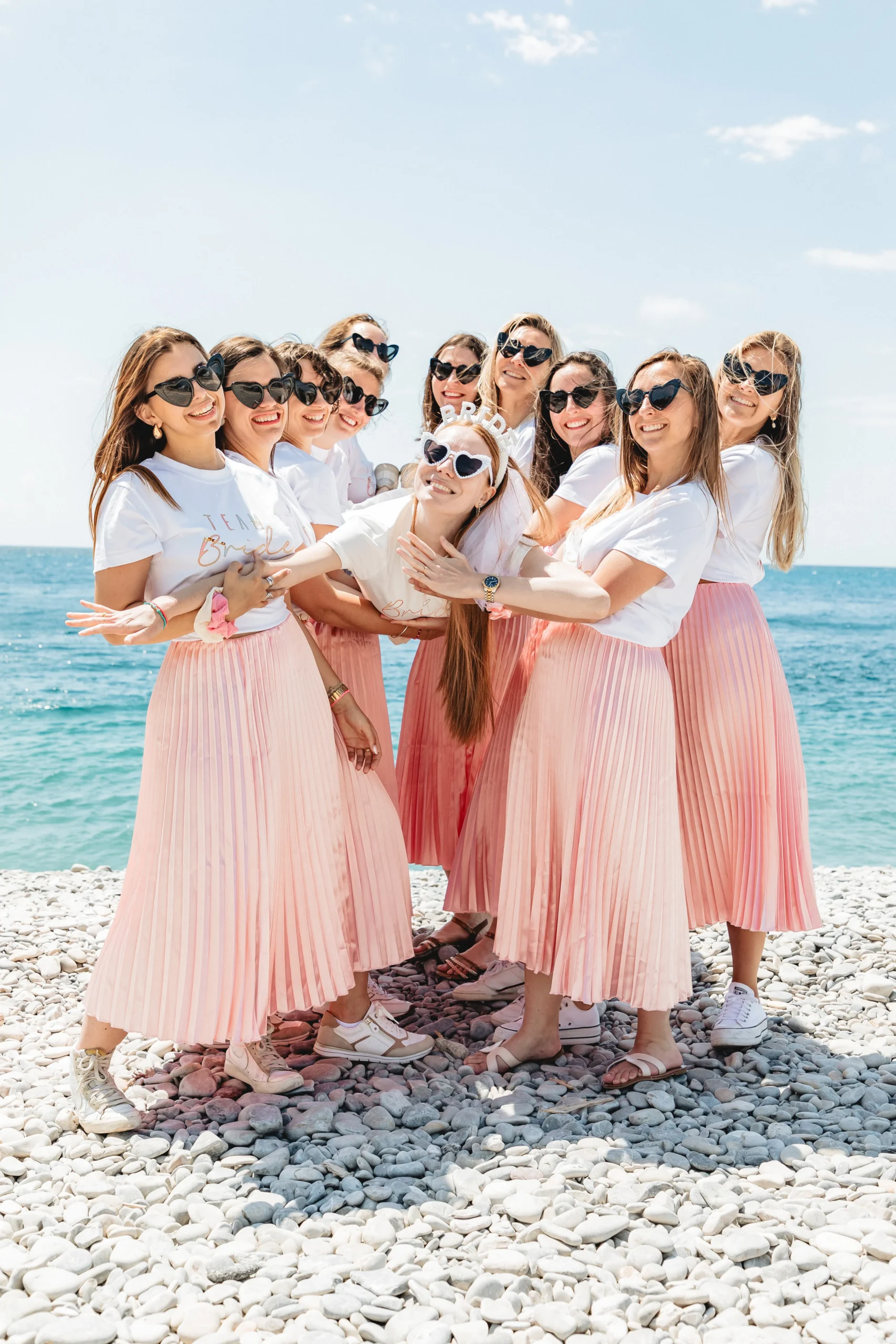 evjf-nice-mariee-porte EVJF à Nice – Les amies soulèvent joyeusement la mariée au-dessus des galets avec la mer en toile de fond