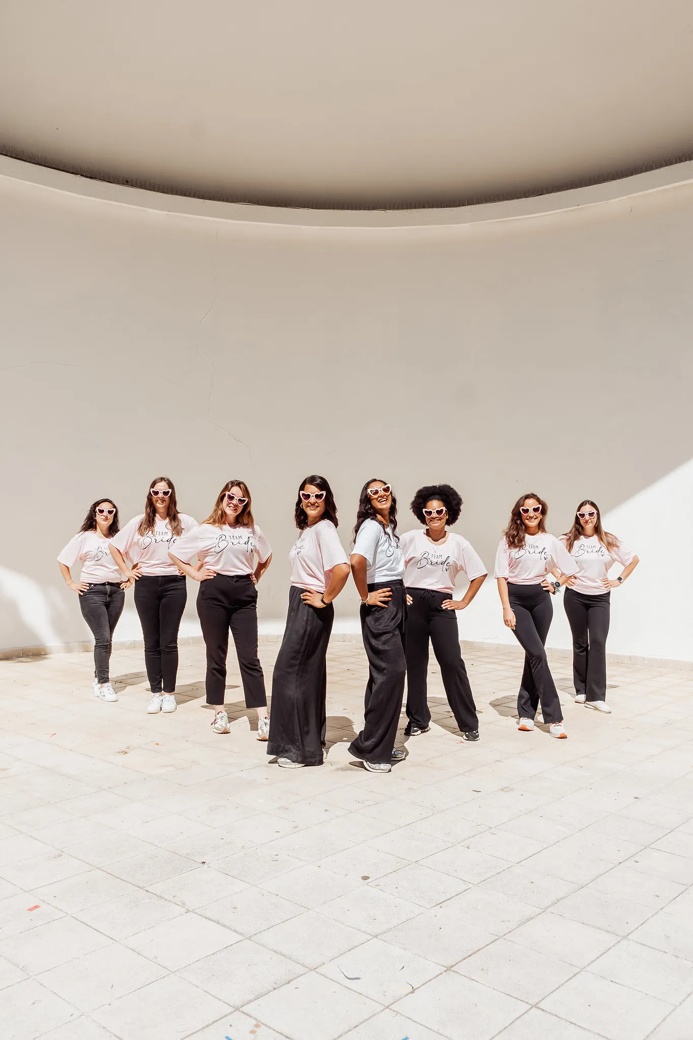 Un groupe de femmes posant en formation, vêtues de tenues assorties “Team Bride”, lunettes en cœur, dans un espace architectural lumineux à Porto.