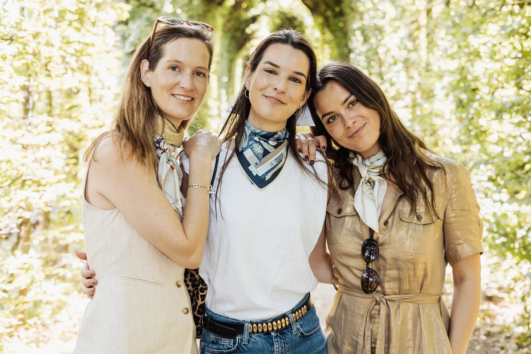 EVJF à Vienne – Portrait chaleureux entre amies dans la verdure Trois femmes souriantes posent ensemble dans un parc arboré pendant un EVJF à Vienne, portant des foulards autour du cou.